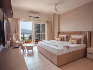 A hotel room with a large bed, a TV, a sofa, and a balcony with a view of buildings and mountains. - Luxury 4 Bedroom's Apartment (Rishikesh)