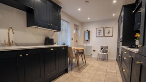 A modern kitchen with dark cabinetry, a central island, and a dining area with two chairs. - The Cove (Lowestoft)
