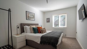 A bedroom with a bed, bedside tables, a radiator, a window with blinds, and a wall-mounted TV. - The Cove (Lowestoft)