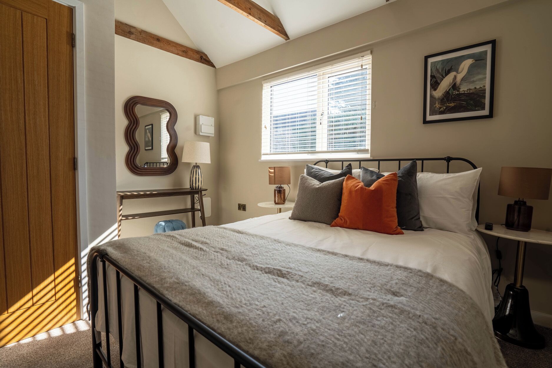 A bedroom with a bed, bedside lamps, a mirror, a window with blinds, and a framed picture on the wall.