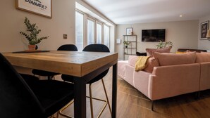 A modern living room with a wooden dining table, two black chairs, a pink sofa, and a TV mounted on the wall. - Coral Cottage (Lowestoft)