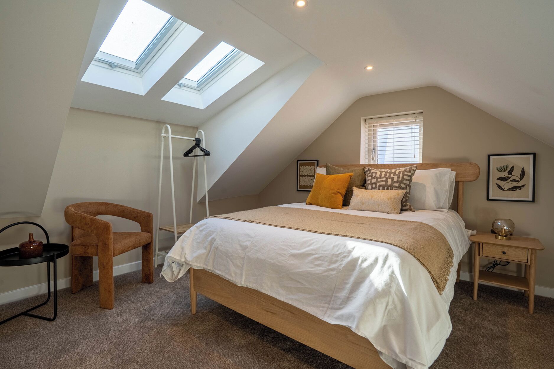A bedroom with a sloped ceiling, skylights, a bed with multiple pillows, a chair, a small table, and a nightstand.