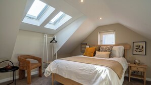 A bedroom with a sloped ceiling, skylights, a bed with multiple pillows, a chair, a small table, and a nightstand. - Coral Cottage (Lowestoft)