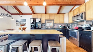 A modern kitchen with wooden cabinets, a large island with stools, stainless steel appliances, and a hanging light fixture.