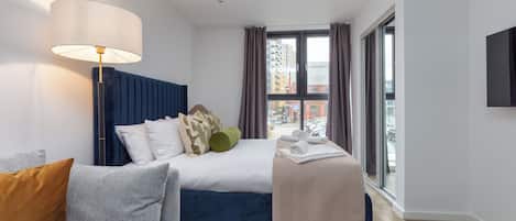 A modern hotel room with a bed, a sofa, a lamp, and a large window offering a city view.