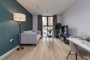 A modern living room with a grey sofa, a floor lamp, a small table, a TV, and a dining area with a white table and chairs. - Cable Street Serviced Apartments (Manchester)
