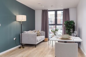A modern living room with a grey sofa, a floor lamp, a dining table set with plates and glasses, and a large plant. - Cable Street Serviced Apartments (Manchester)