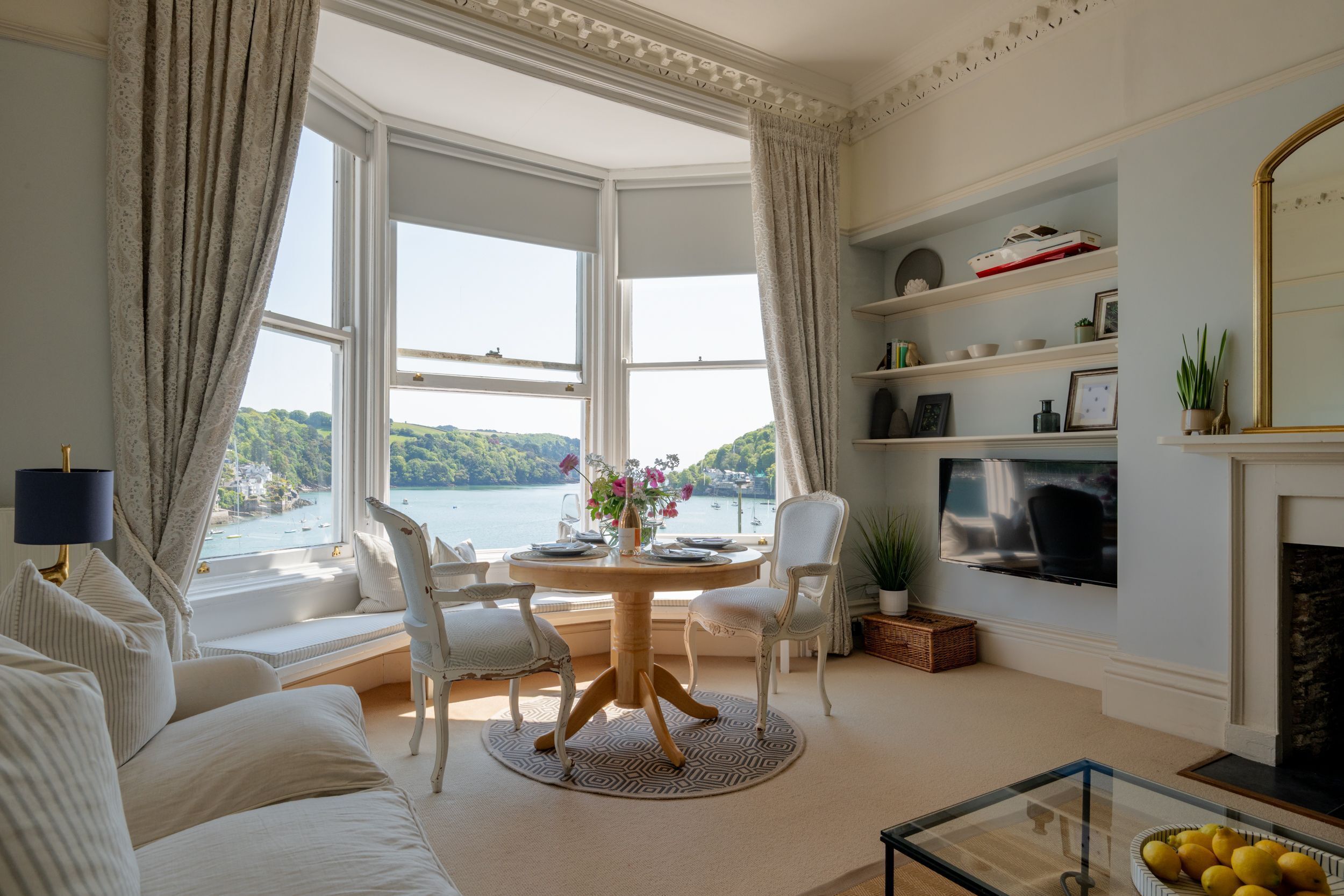 A living room with a bay window overlooking a body of water, a fireplace, and a television.