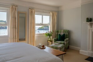 A bedroom with a large bed, a chair with a green cushion, a vase with flowers, and a view of the sea through the window.