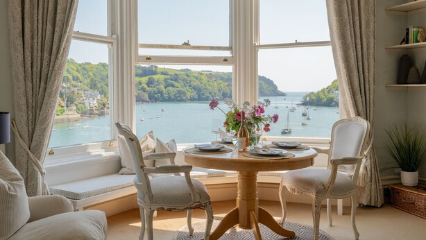 A room with a bay window overlooking a body of water, a dining table set with plates, and two chairs.