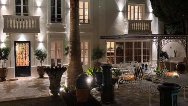 Un edificio blanco con múltiples ventanas y balcones, rodeado de plantas en macetas y mesas al aire libre.
