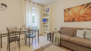 Een woonkamer met een bank, een eettafel met stoelen, een boekenkast, en een groot bloemenstillevaan aan de muur.