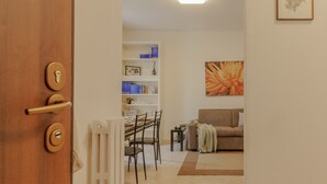 Een kamer met een houten deur, een witte radiator, een eettafel met stoelen, een bank en een boekenkast met diverse spullen.