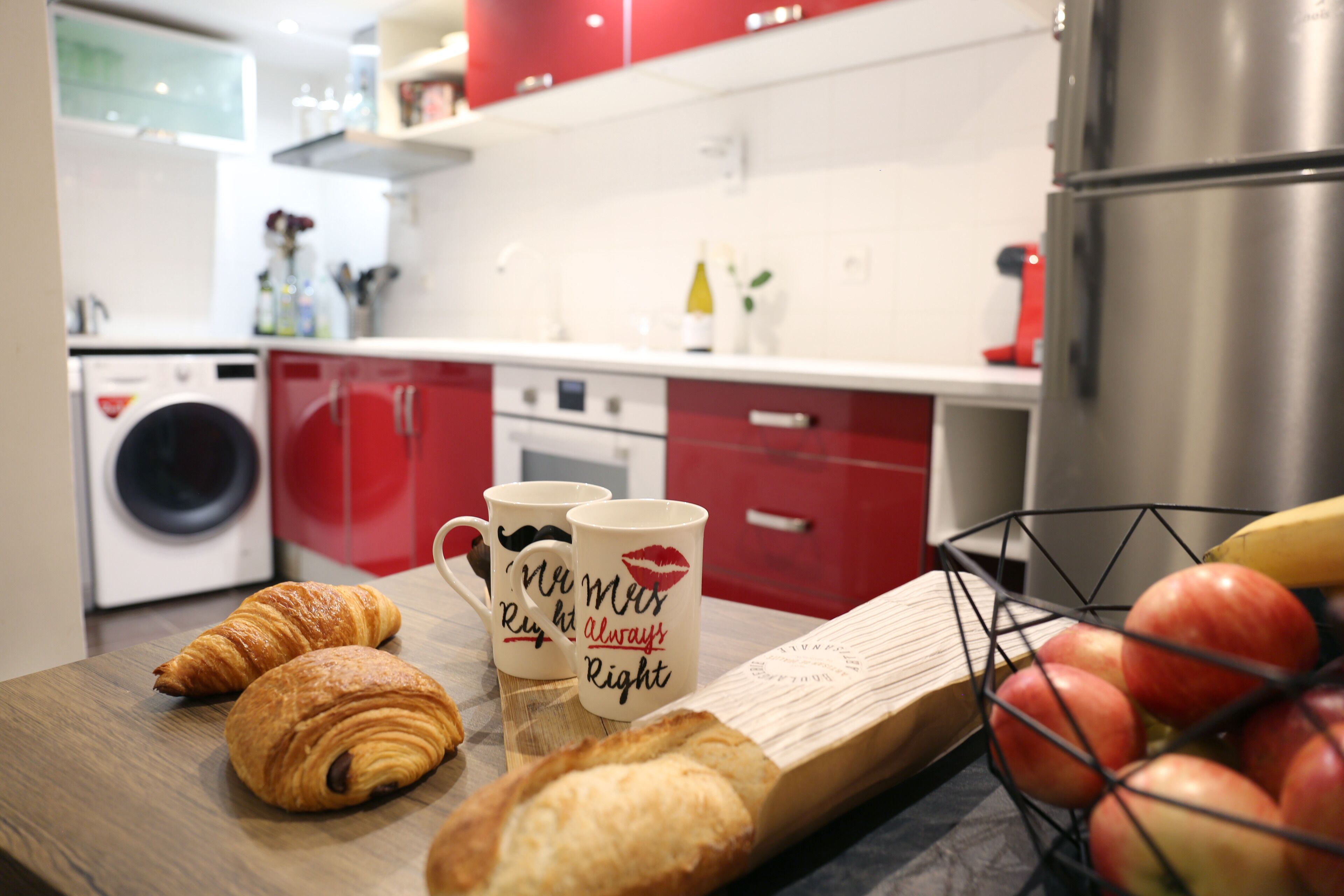A kitchen with red cabinets, a washing machine, a coffee maker, and a fruit basket.