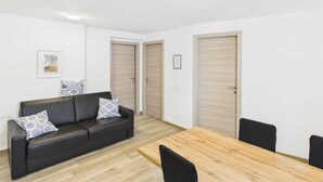 Un salon moderne avec un canapé en cuir noir, des coussins à motifs, une table à manger en bois et deux portes en bois.