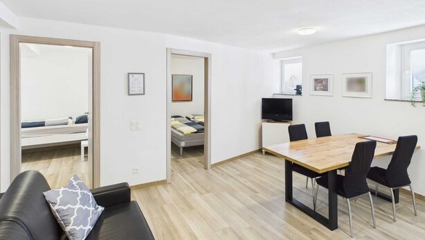 A modern, well-lit room with a wooden dining table, black chairs, a black sofa with a patterned pillow, a television, and a bed in the adjacent room. - 1 Curio Malcantone Charming Lodge (Curio)