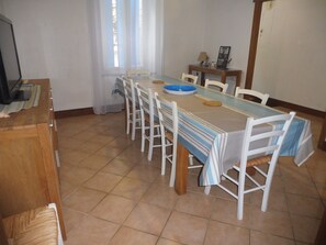 A dining area with a long table, white chairs, a blue and white striped tablecloth, a blue bowl, and a small table with a lamp and picture frame.