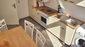 Cocina compacta con encimera de madera, microondas, horno, lavaplatos y lavadora. Hay sillas blancas y una mesa.