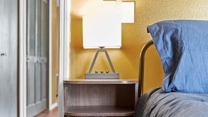A bedroom with a wooden nightstand, a lamp, a bed with blue bedding, and a wooden floor. - Joplin Avenue Junction: Two Bedroom Duplex (Tulsa)