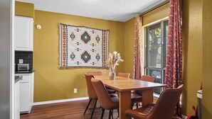 A dining area with a wooden table and chairs, a patterned wall hanging, and a window with curtains. - Joplin Avenue Junction: Two Bedroom Duplex (Tulsa)