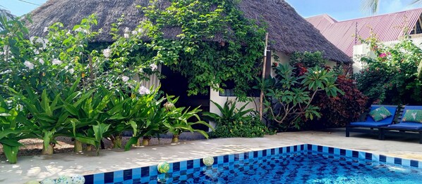 A pool area with a tiled edge, surrounded by lush greenery and a thatched-roof structure. - Aurora Boutique Hotel (Jambiani)