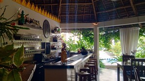 A bar area with a thatched roof, shelves with bottles, glasses, and a large decorative plate. There are wooden stools and a view of a pool outside. - Aurora Boutique Hotel (Jambiani)
