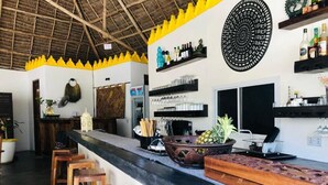 A bar area with wooden stools, a black countertop, and shelves with bottles and glasses. - Aurora Boutique Hotel (Jambiani)