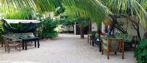 Un espacio al aire libre con mobiliario de mimbre y suelo de grava, rodeado de abundante vegetación y palmeras.