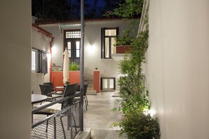 An outdoor patio area with tables and chairs, a small garden, and a staircase leading to an upper level. - Vol3 (Athens)