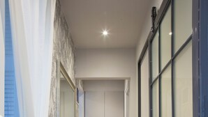 A hallway with wooden flooring, a white door at the end, and a large window with a view of a building. - Vol3 (Athens)