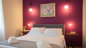 A neatly made bed with white linens and a grey blanket, a bedside table with a vase of flowers, and a wall-mounted painting of flowers. - Vol3 (Athens)