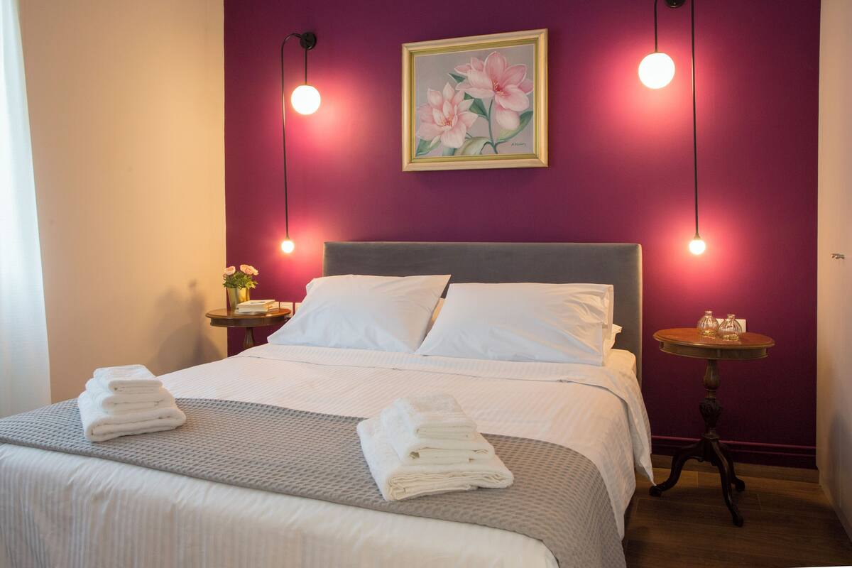 Ein ordentlich bezogenes Bett mit weißen Leinen und einer grauen Decke, ein Nachttisch mit Blumen in einer Vase und ein an der Wand hängendes Blumenbild.