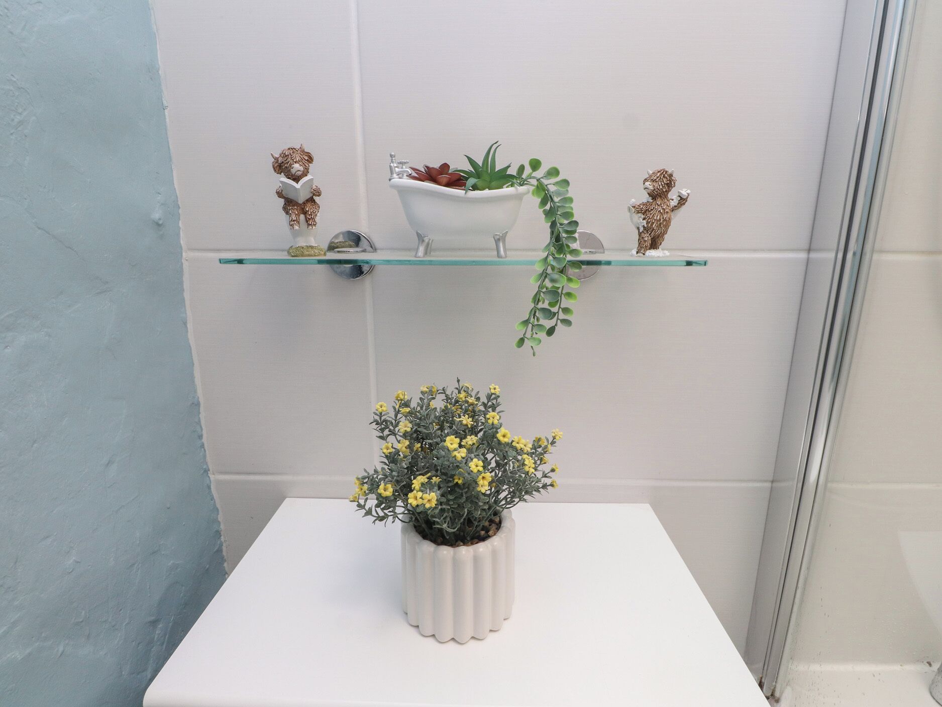 Een badkamer met een wit wastafel, een glazen plank met decoratieve voorwerpen, en een potplant op een witte tafel.