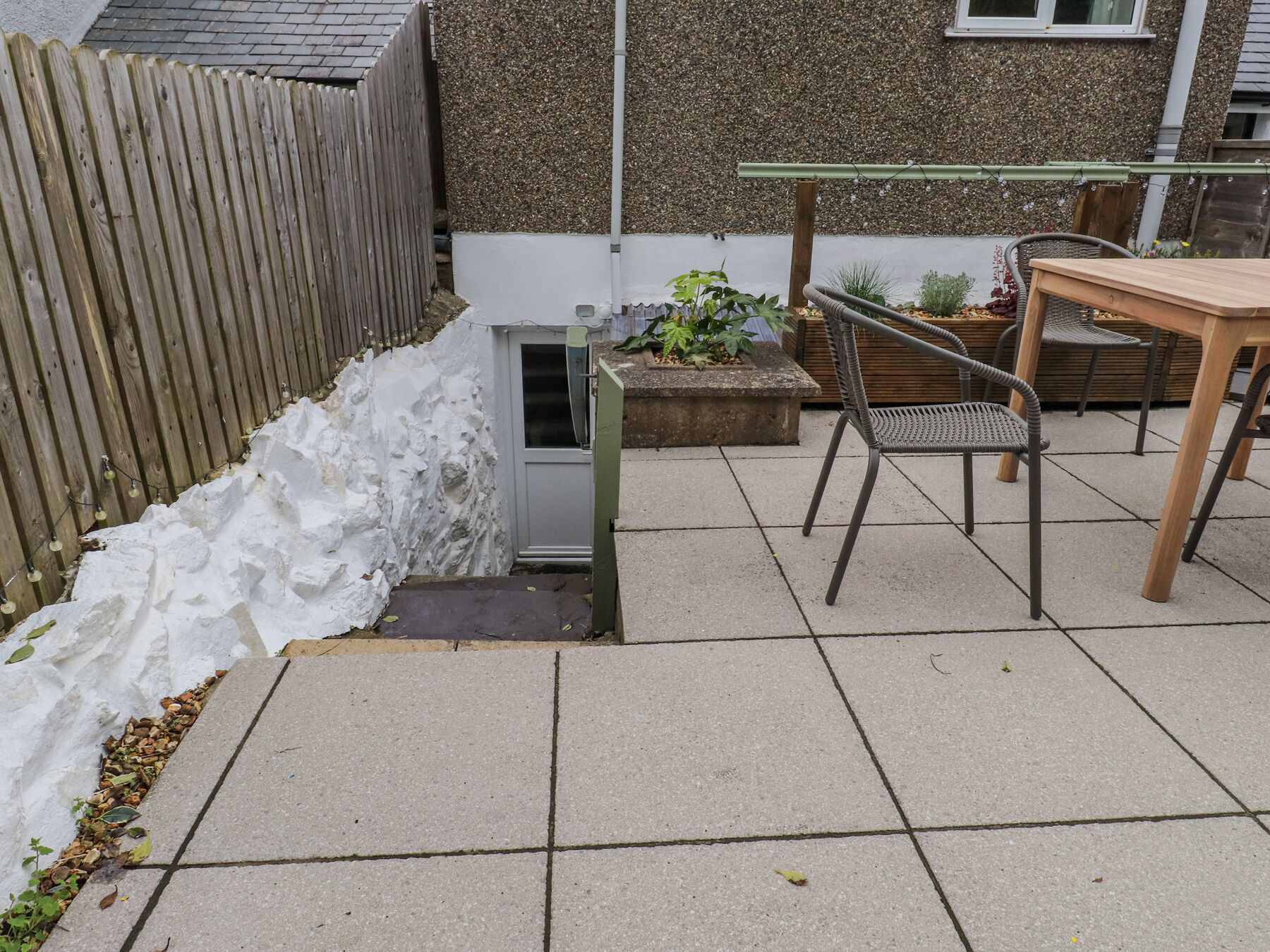 A patio area with a wooden table and chairs, a small garden bed with plants, and a white wall with a window.