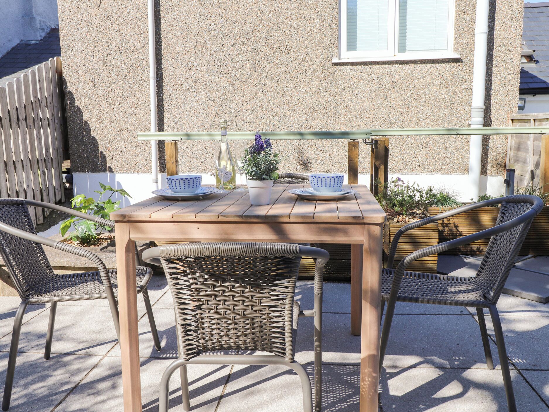 An outdoor seating area with a wooden table and wicker chairs, a bottle of wine, and a bowl on the table.