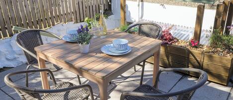 Una mesa de madera preparada para cuatro personas, con sillas de mimbre, rodeada por una cerca de madera y un parterre con flores.