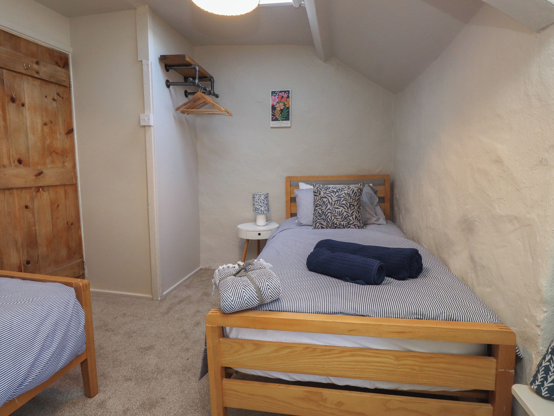 A small, cozy bedroom with a wooden bed, a bedside table, a hanging coat rack, and a small wall decoration.