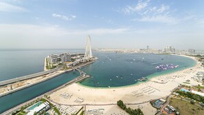Un paysage urbain côtier avec une grande roue bien visible, une plage parsemée de parasols et un port de plaisance rempli de bateaux.