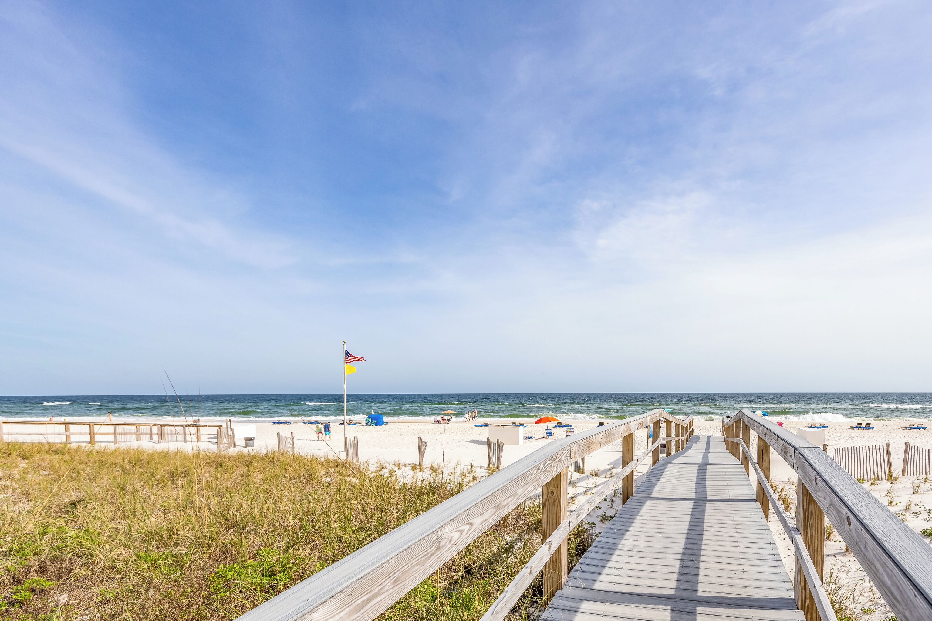 A wooden boardwalk leading to a beach with a flag, sandy shore, and ocean view.
