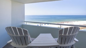 Un balcón con dos sillas Adirondack y una mesa, con vistas a una playa con sombrillas y una piscina.