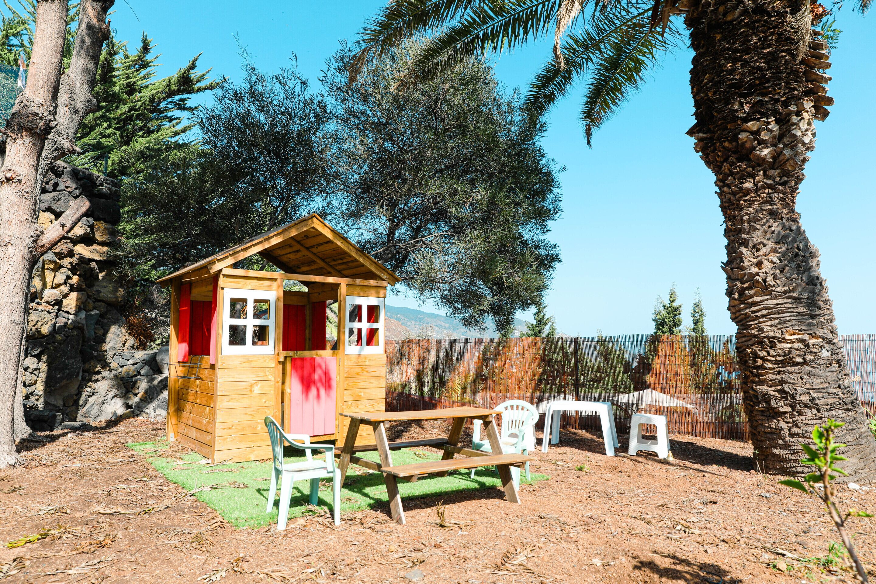 Une cabane en bois avec une porte rouge, une table de pique-nique et deux chaises en plastique blanches, dans un jardin avec des arbres et un mur de pierres.