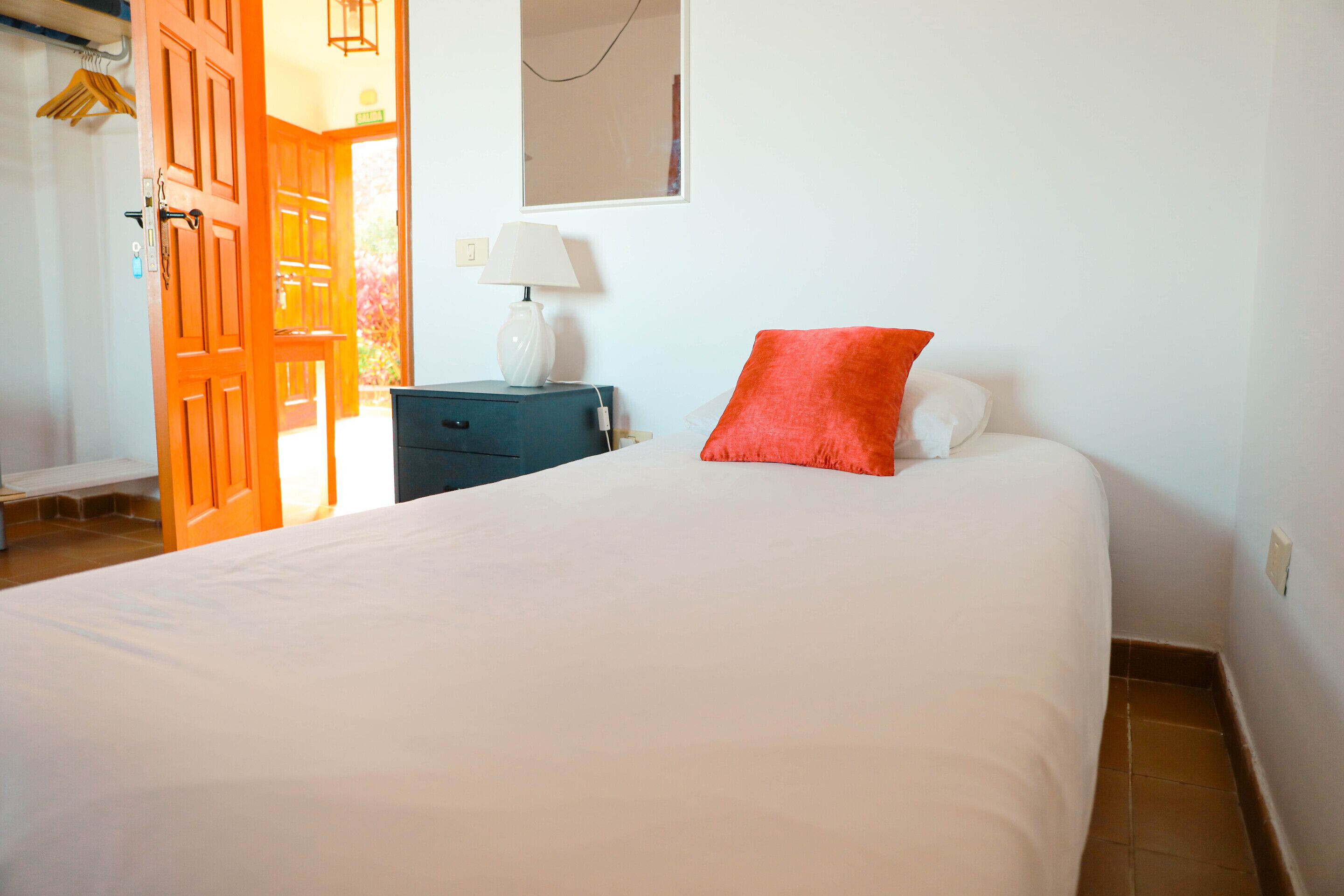 A neatly made bed with a white comforter and a red pillow, a small bedside table with a lamp, and a wooden door leading to another room.