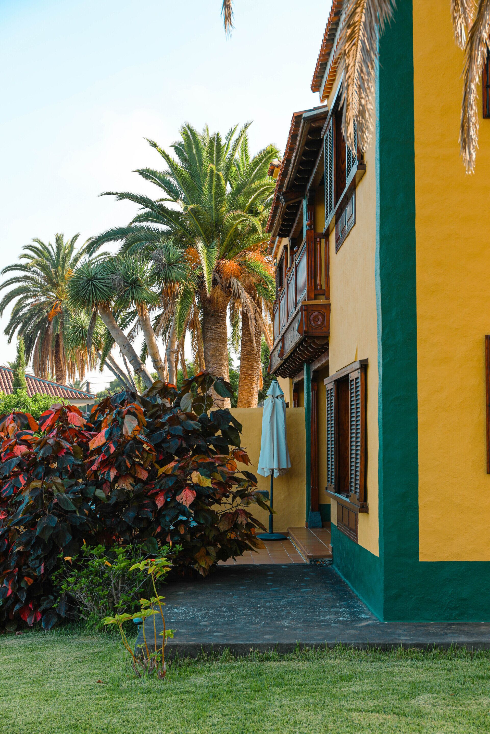 A two-story building with a green and yellow facade, palm trees, and a small garden area with a bench and umbrella.