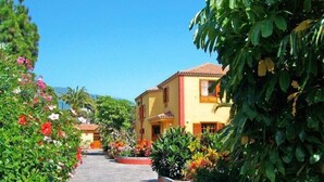 Un sendero conduce a un edificio amarillo con techo rojo, rodeado de abundante follaje y plantas en flor.