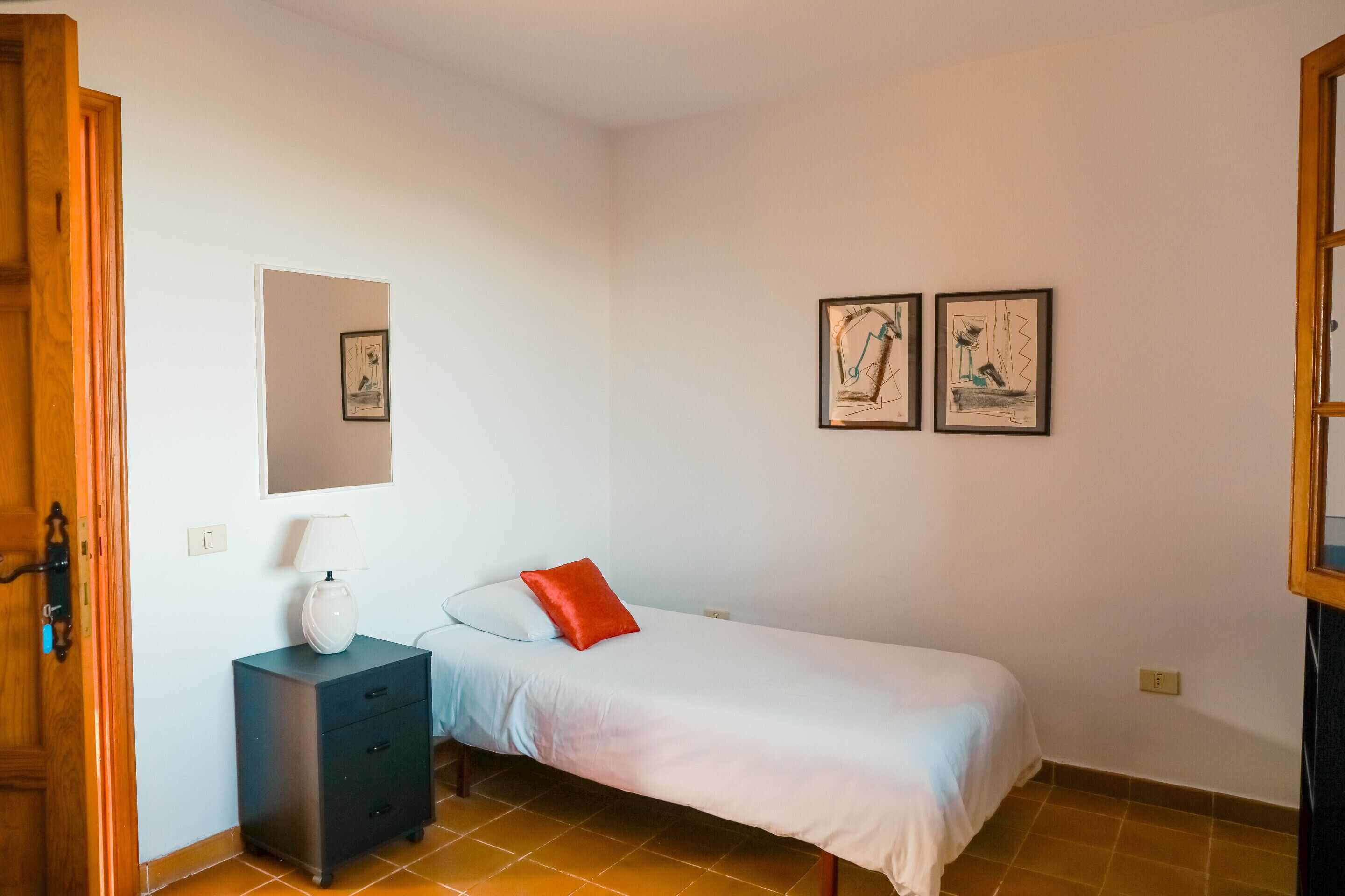 Una habitación pequeña con una cama individual, una mesita de noche, una lámpara y dos cuadros en la pared.