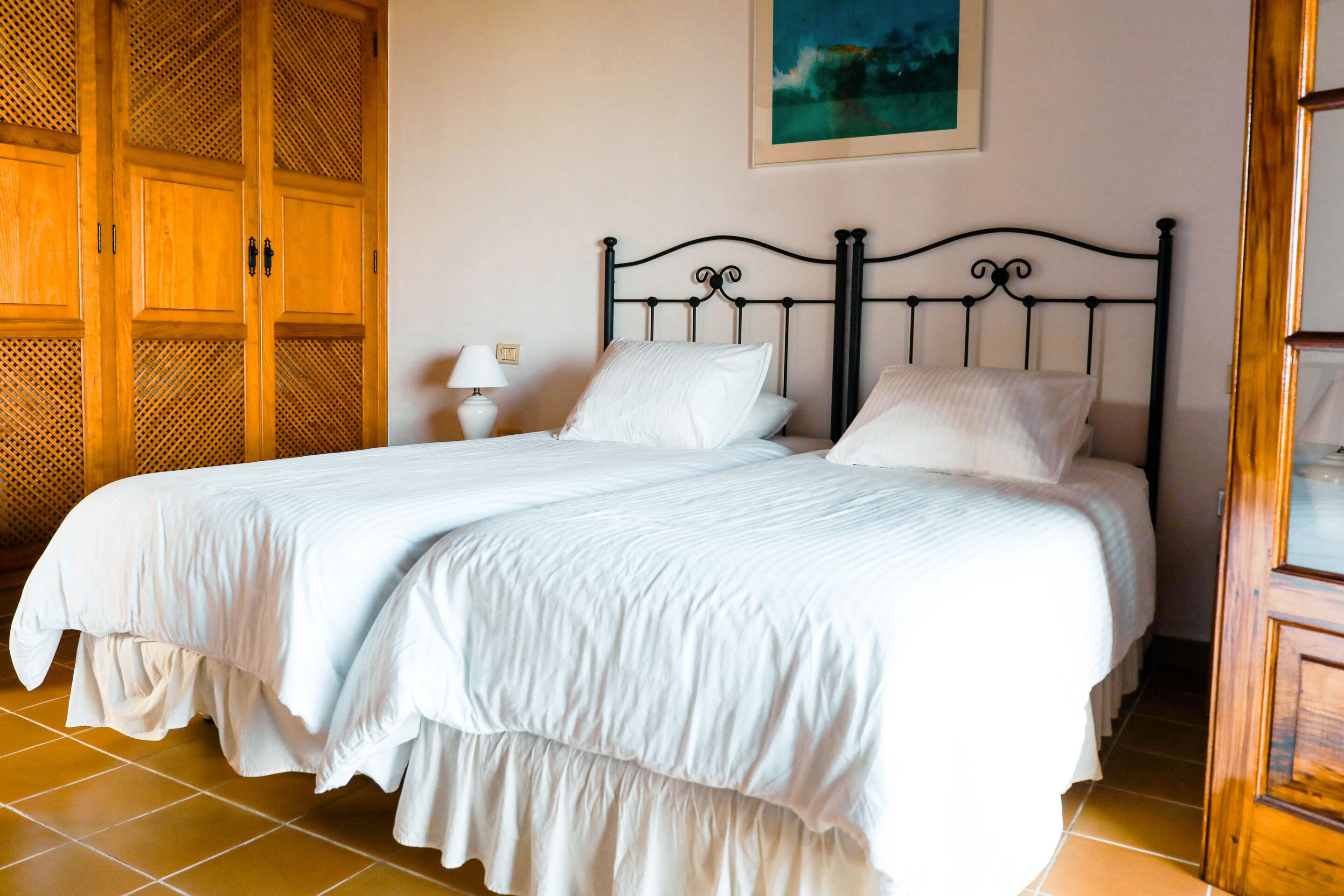 A neatly made bed with white linens, a wooden wardrobe with louvered doors, and a painting on the wall.