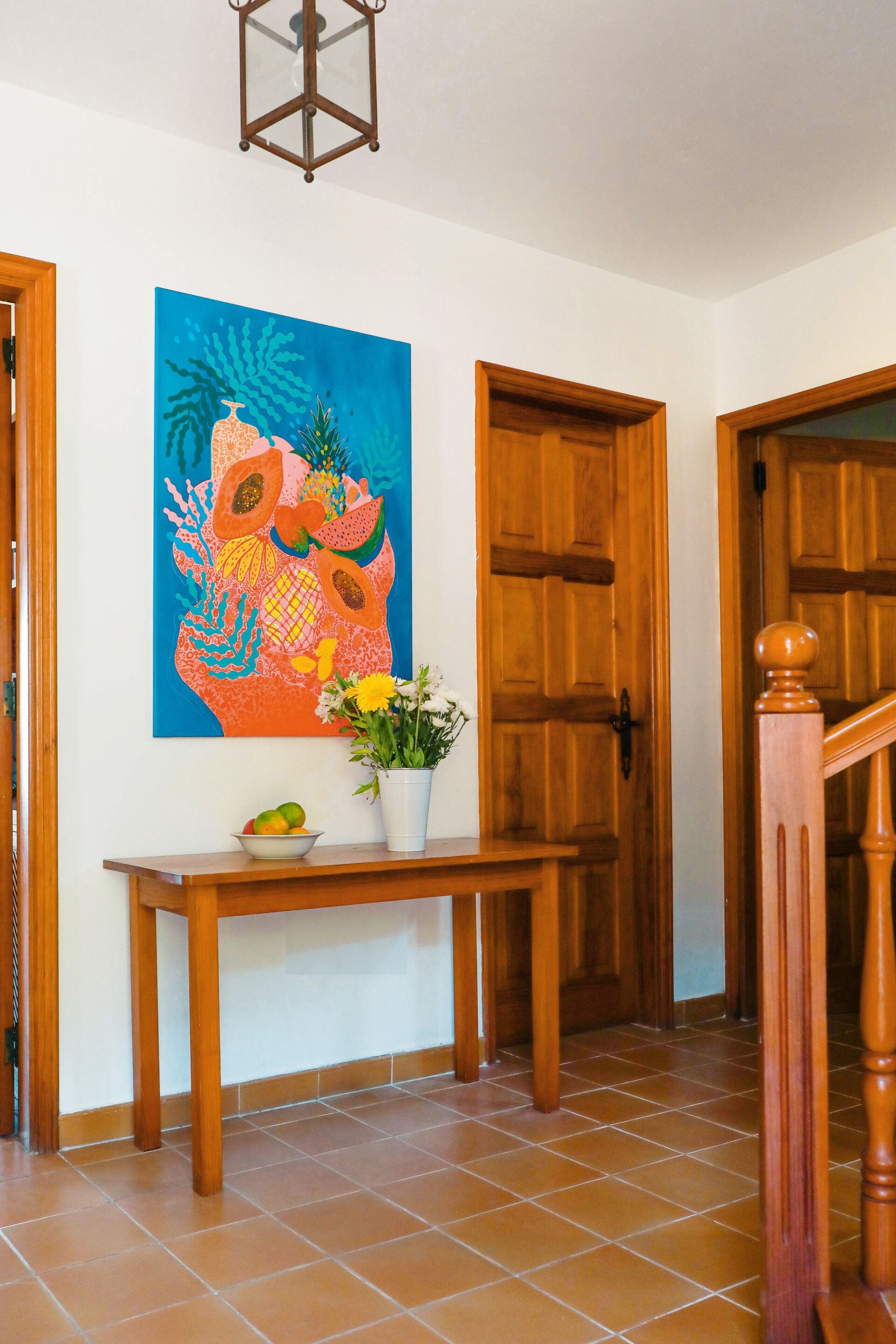 Un pasillo con puertas de madera, piso de baldosas, una consola de madera con un florero y un cuadro de colores en la pared.