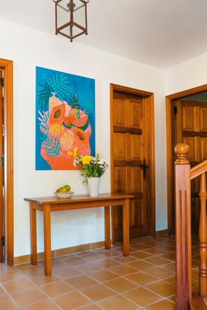 A hallway with wooden doors, a tiled floor, a wooden console table with a vase of flowers, and a colorful painting on the wall.