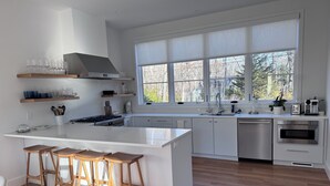 A modern kitchen with a central island, wooden stools, a large window, and stainless steel appliances. - New construction, 4-bedroom house with, fitness room in lovely Bridgehampton (Bridgehampton)