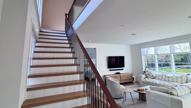 A modern staircase with wooden steps and black railings, leading to a living area with a sofa and a television. - New construction, 4-bedroom house with, fitness room in lovely Bridgehampton (Bridgehampton)